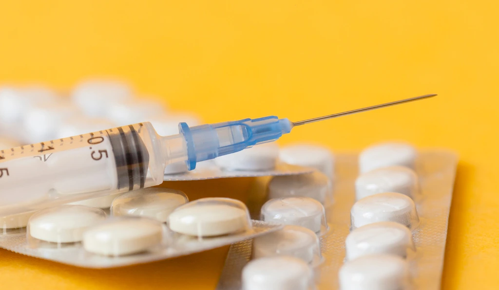 A close-up image of a syringe with a blue needle cap resting on top of multiple blister packs of white pills, set against a yellow background.